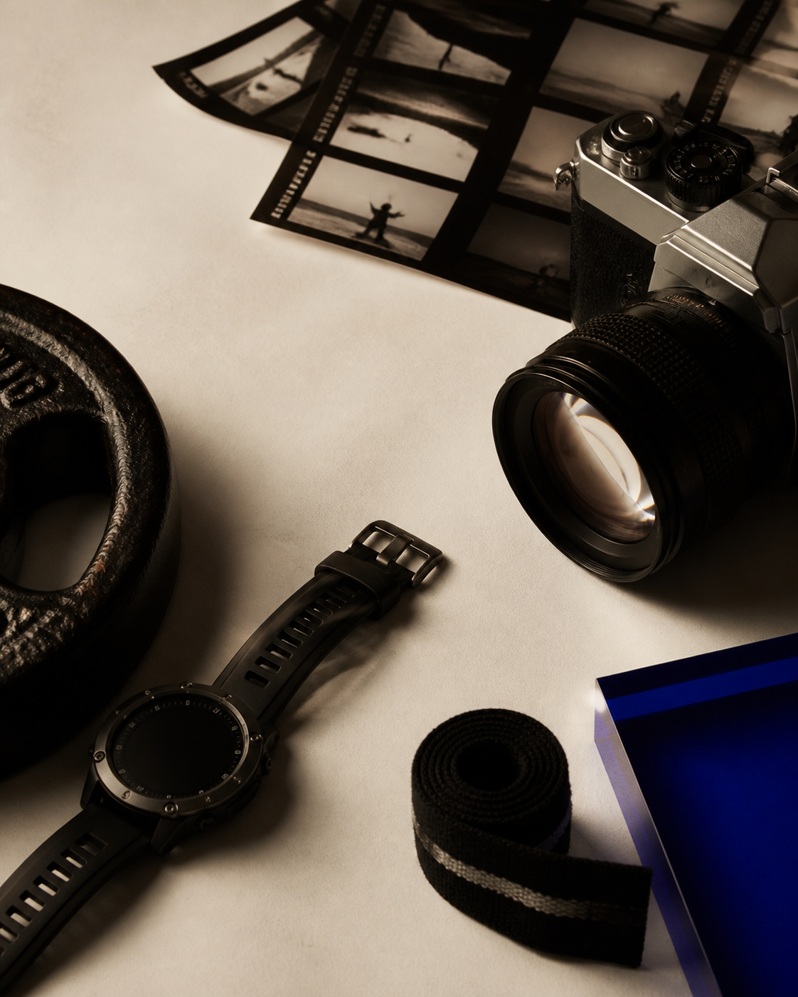 A camera, training watch, weight, wrist wrap and contact sheets on a warm ivory surface